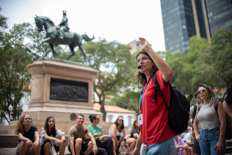 Guia falando enquanto o grupo está sentado ouvindo. Ao fundo, é possível ver a estátua do General Osório.